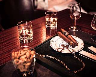 Two cigars on a plate beside whisky glasses and a cognac glass on a wooden table.