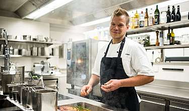 Un cuisinier se tient dans une grande cuisine moderne à côté d’un plat à rôtir chaud contenant de la viande.