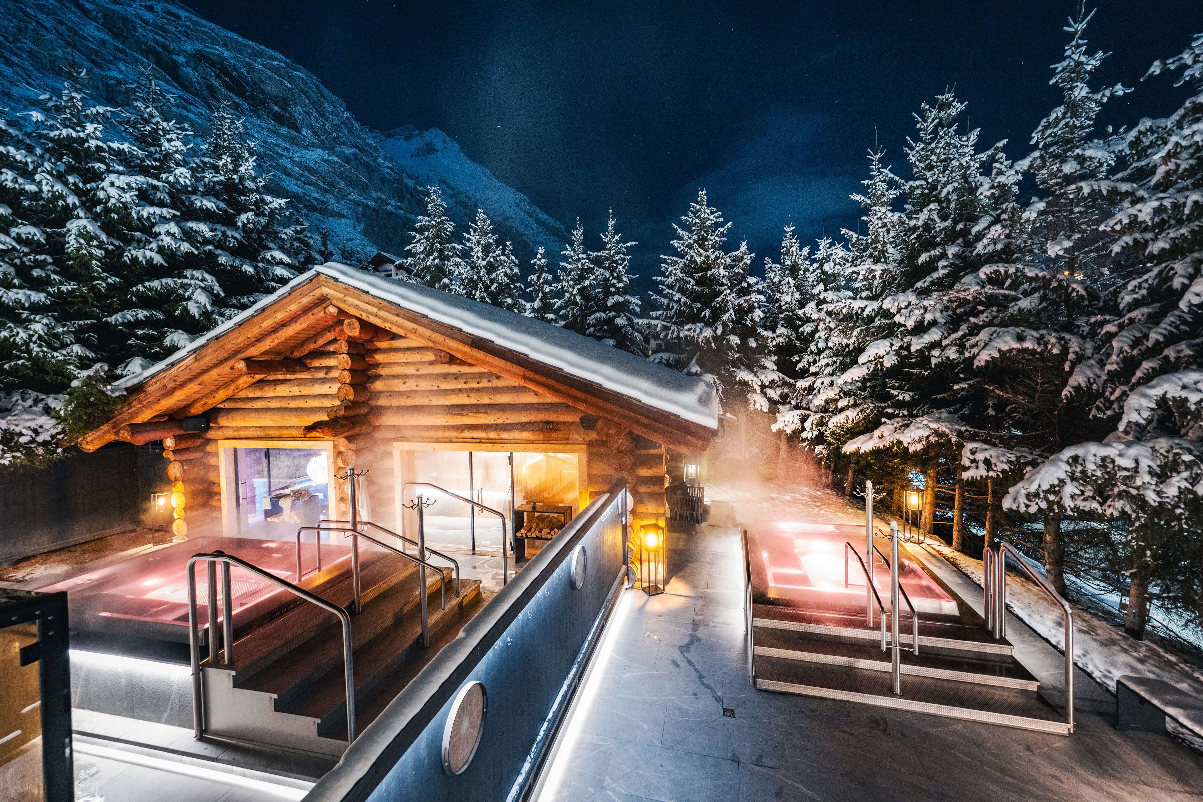Beleuchtete Saunahütte mit zwei Außen-Whirlpools in einer verschneiten Berglandschaft bei Nacht.