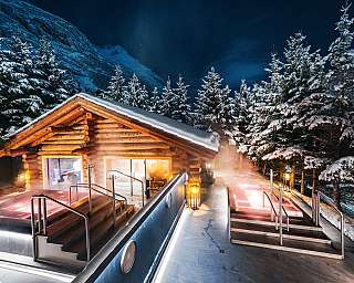 Wooden chalet in the snow with illuminated outdoor whirlpools between snow-covered fir trees at night.