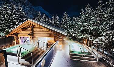 Chalet en bois avec des piscines extérieures éclairées dans un paysage de montagne enneigé la nuit.