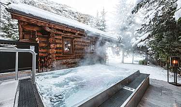 Cabane en bois dans la neige à côté d'un bain à remous extérieur fumant dans une forêt hivernale.
