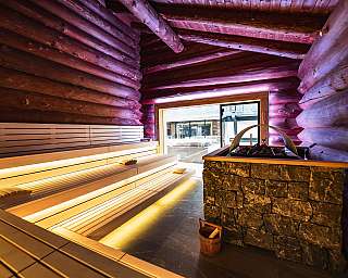 Innenraum einer Sauna mit Holzbanken, Steinofen und hell beleuchteter Holzverkleidung.
