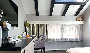 Helles Dachzimmer mit sichtbaren Holzbalken, Schreibtisch, Stuhl und großen Fenstern.