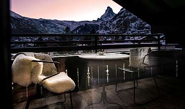 Balcon confortable avec table et chaises devant des montagnes enneigées au coucher du soleil.
