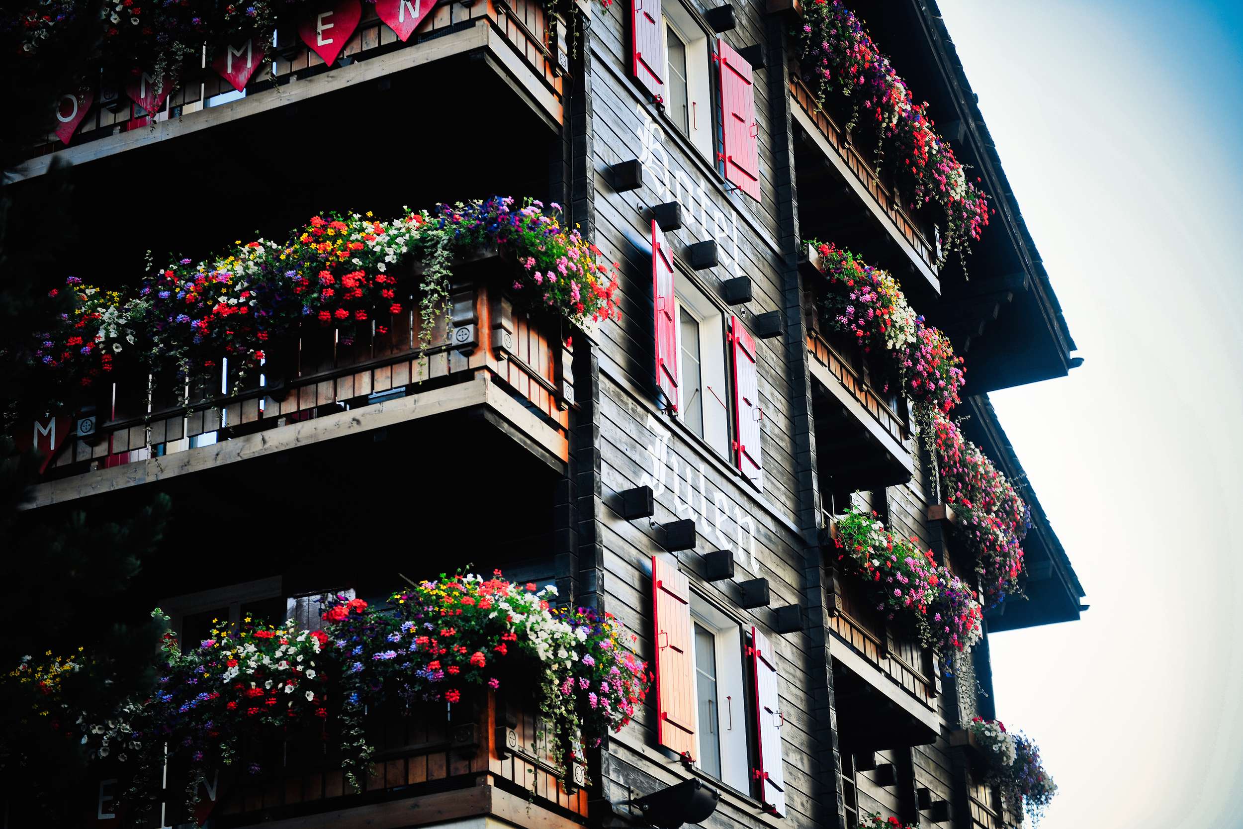 Maison en bois à plusieurs étages avec des balcons abondamment fleuris et des volets rouges.
