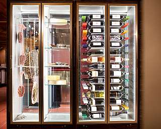 Beleuchteter Kühlschrank mit Weinflaschen, Käserädern und hängenden Wurstwaren.