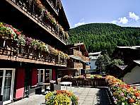 Maison en bois avec balcons décorés de fleurs devant une montagne boisée et un ciel bleu.