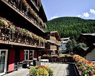 Holzhotel mit blühenden Balkonen vor bewaldetem Berg und blauem Himmel.