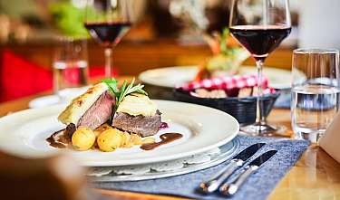 Serviertes Fleischgericht mit Kartoffeln, Sauce, Rotwein und Wassergläsern auf einem gedeckten Tisch.