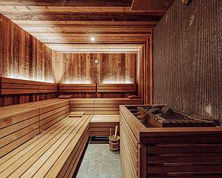 Sauna moderne avec bancs en bois, poêle avec pierres et murs éclairés par une lumière chaude.