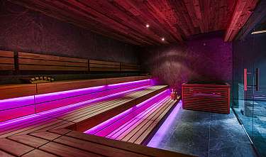 Sauna en bois avec éclairage LED violet, paroi en verre et sol sombre en pierre.
