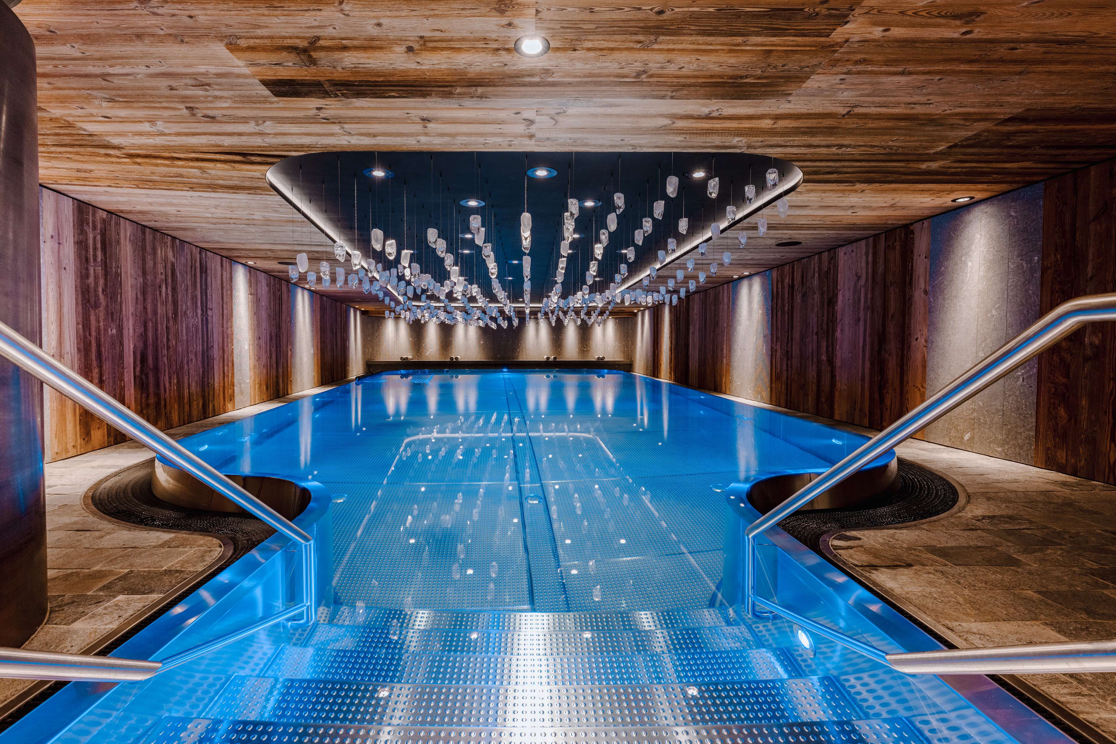 Piscine intérieure moderne avec une surface d’eau bleue, un plafond en bois et des suspensions en verre.