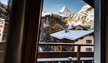 Chalets enneigés devant un sommet de montagne remarquable, vus à travers une fenêtre.