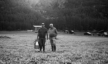 Zwei Männer stehen auf einer Wiese vor bewaldeten Bergen und kleinen Hütten.
