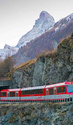 Un train de montagne rouge longe une paroi rocheuse devant des Alpes enneigées.