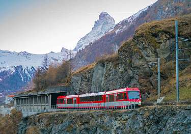 Un train rouge traverse un paysage alpin rocheux avec des montagnes enneigées.