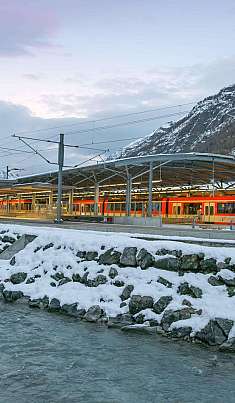 Train rouge dans une gare d’une vallée de montagne enneigée au bord d’une rivière.