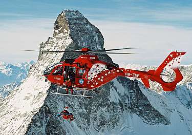 Hélicoptère rouge volant devant des sommets enneigés avec un sportif en rappel.