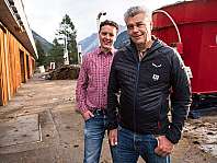 Deux hommes se tiennent devant une installation rouge à côté d’un bâtiment en bois dans un paysage de montagne.