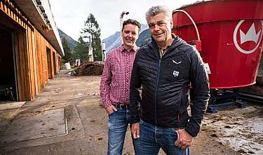 Zwei Männer stehen vor einem Gebäude und einem roten Industriebehälter in einer Berglandschaft.