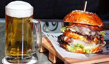 A glass of beer and a large burger on a wooden table.