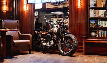 Vintage-Motorrad steht in einem holzgetäfelten Raum neben einem Ledersessel und Bücherregalen.