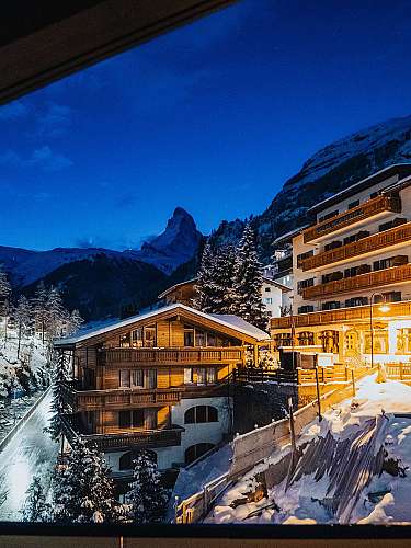 Beleuchtete Alpenhäuser an einem verschneiten Fluss unter blauem Abendhimmel.