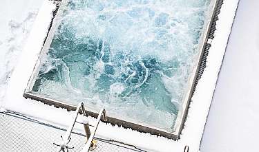 Ein mit Schnee bedeckter Whirlpool mit sprudelndem, blauem Wasser und Leiter an Deck eines Schiffes oder Boots.