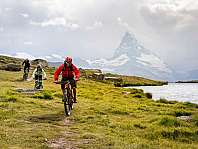 Trois vététistes roulent sur un chemin herbeux le long d’un lac, devant une montagne enneigée.
