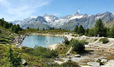 Bergsee in alpiner Landschaft mit Wanderern, Bäumen und schneebedeckten Gipfeln.