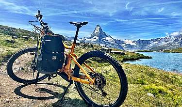 Ein Mountainbike steht auf einem Pfad vor Bergsee und schneebedecktem Gipfel.