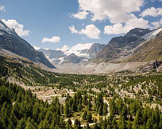Vaste vallée de montagne avec forêt de conifères, parois rocheuses abruptes et sommets enneigés sous un ciel bleu.