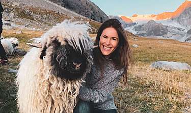 Lachende Frau kniet neben einem großen Bergschaf in einer alpinen Landschaft.