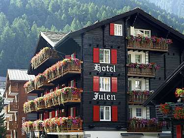 Hôtel de montagne traditionnel avec volets rouges et balcons fleuris devant des montagnes boisées.