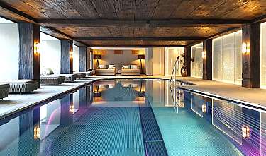 Piscine intérieure moderne avec plafond en bois, chaises longues, canapés et murs éclairés d’une lumière chaleureuse.