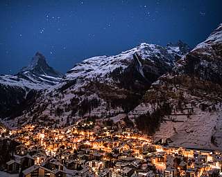 Lights of a snow village beneath rocky, snow-covered mountains under a starry sky.