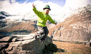 A child in a green jacket is jumping on a rock in front of a mountain landscape.