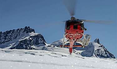 Roter Helikopter schwebt vor verschneiten Berggipfeln.