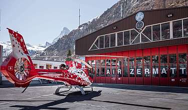 Roter Helikopter steht vor einem Hangar mit Bergkulisse.