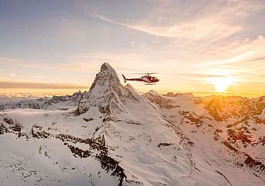 Roter Hubschrauber fliegt bei Sonnenuntergang über verschneite Berggipfel.
