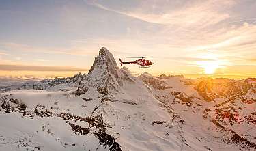 Roter Hubschrauber fliegt bei Sonnenuntergang über eine verschneite Berglandschaft.