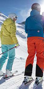 Zwei Skifahrer stehen auf einer verschneiten Piste vor einem Berg im Sonnenlicht.