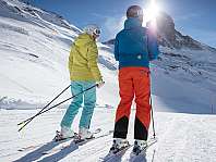 Deux skieurs se tiennent sur une piste enneigée devant une montagne au soleil.