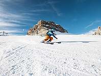 Un skieur descend une large piste enneigée devant des montagnes rocheuses.