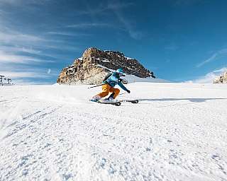 Un skieur descend une piste enneigée devant des montagnes rocheuses.