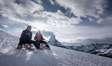Zwei Personen sitzen auf Schlitten in einer verschneiten Berglandschaft.