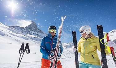Deux skieurs se tiennent avec des skis et des bâtons de ski dans un paysage de montagne ensoleillé et enneigé.