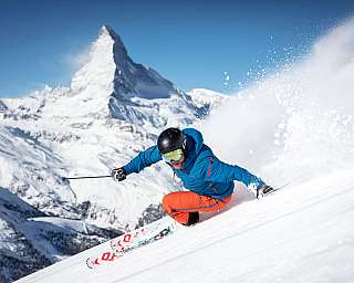 A skier in a blue jacket and orange trousers skiing down a steep, snowy piste.