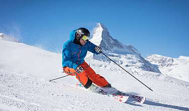 Un skieur en veste bleue et pantalon orange descend dans la neige devant un décor de montagne.